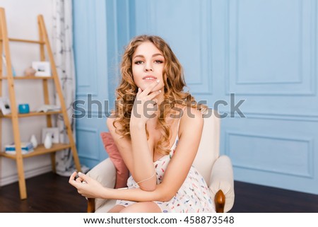 Hot stylish girl with blond curly hair sits on armchair in studio with blue walls and brown furniture. She wears white dress. She listens to music on smartphone.