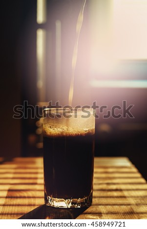 Milk pouring in transparent glass with coffee drink. Kitchen table in sun light. Image toned, lens flare effect added. Good morning concept.