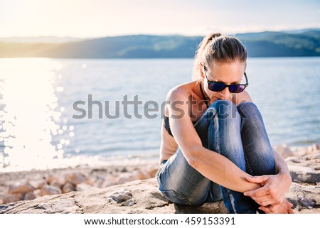 Sad woman with sunglasses wearing black bikini and jeans sitting on the stone dock and thinking Royalty-Free Stock Photo #459153391
