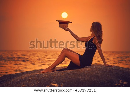 Young woman sits on the rock and hold a hat under the sun during sunset