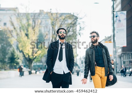 two young bearded blonde and black hair modern businessman, walking in the city backlight, one overlooking right, the other overlooking left - working, successful concept