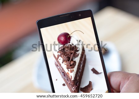 Closeup of hands making photo of sweet dessert on mobile phone while sitting in restaurant, and  focus at the chocolate on topping of a sliced black forest cake