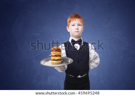 Little waiter stands with tray serving big double hamburger. Redhead Child boy in suit plays restaurant servant, gives burger at blue background