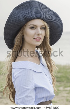 portrait of a cute blonde long-haired caucasian woman in a blue shirt standing by the water smiling holding a hat on her head