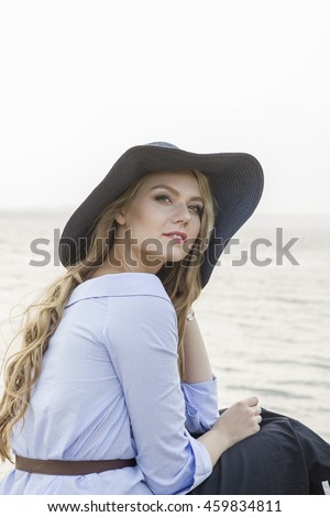 cute blonde long-haired caucasian woman sitting by the water with a hat on her head