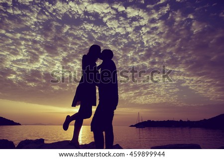 Silhouette of a couple in love hugging and kissing in Syvota port, Greece in sunset. Sunset, sunlight, Ionian sea and cloudy sky in the background.