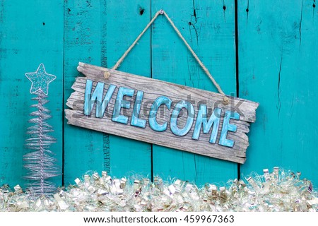 Welcome sign hanging on rustic teal blue wood background with white Christmas garland border and silver tree
