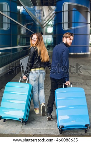 Cute couple is walking with suitcases outside in airport. She has long hair, glasses,  yellow sweater, jacket. He wears black shirt, beard. View from back.
