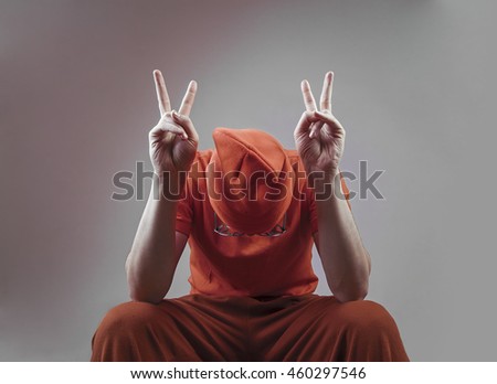 Portrait of male prisoner in orange uniform isolated on gray background. Peace brother - fingers sign. depressed criminal sitting behind bars