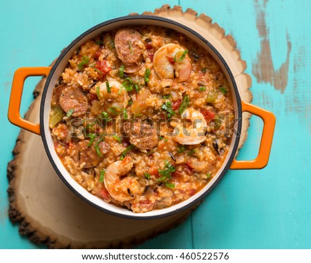 Overhead shot of Jambalaya with scallion garnish in an orange stock pot. Shown on a rustic wooden trivet against an antique turquoise table. 