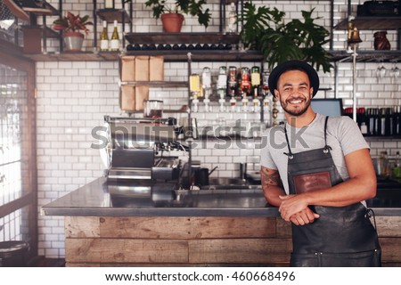 Portrait of young man standing at the counter in his cafe. Coffee shop working in apron and hat smiling at camera. Royalty-Free Stock Photo #460668496