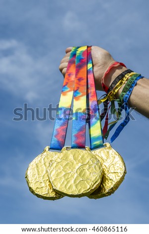 Hand of athlete wearing good luck Brazilian wish ribbons holding gold medals hanging from colorful ribbons against bright blue sky