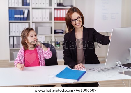 Young businesswoman amusing her small daughter as she babysits her in the office smiling as she shows her something on the computer screen