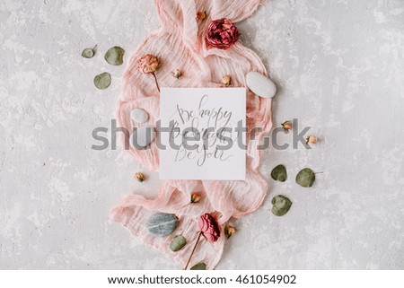 inspirational quote "be happy be bright be you" written in calligraphy style on paper with dry white tulips, eucalyptus petals and pink textile on concrete background. Flat lay, top view