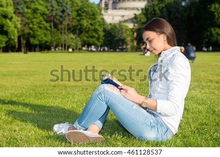 Smiling beautiful young woman on grass and reading book, against background of summer green park.