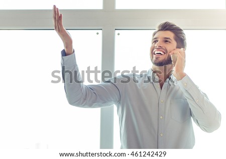 Handsome young businessman in classic shirt is talking on the mobile phone and smiling, looking and pointing upward, standing in office before the window