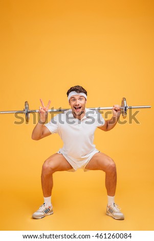 Funny cheerful young sportsman doing squats with barbell and showing peace sign over yellow background
