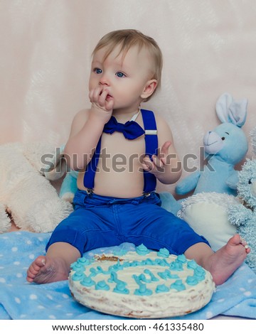 Funny baby boy is tasting the cake and blue butter icing on his sticky fingers from his first birthday cake smash while sitting on a pink and blue background. Toddler sucking fingers with cake.