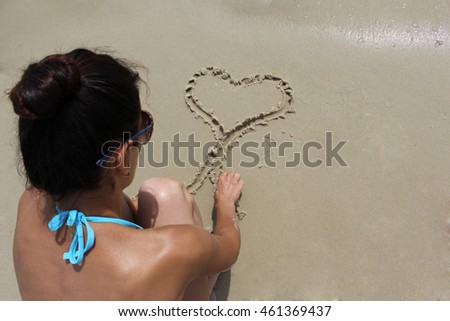 Tanned dark haired girl draws in the sand near the sea. She draws a heart