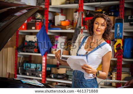beautiful smiling woman mechanic in blue denim overalls with a laptop and sheets of paper in the interior of an automobile repair shop near the car with an open hood