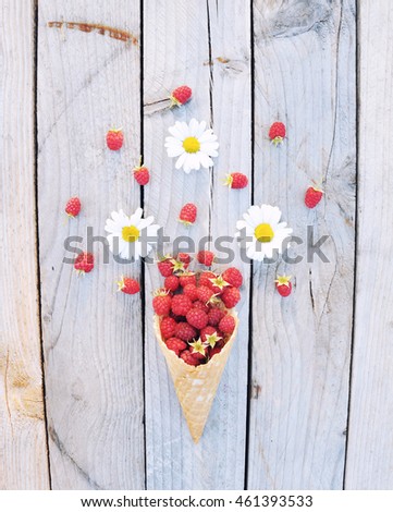 Ripe fresh raspberries and white chamomile flowers in ice cream cone on rustic wooden background. Stylish flat lay. Minimal concept.