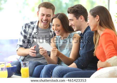 Four casual happy friends laughing watching media content together in a tablet sitting on a sofa in the living room at home