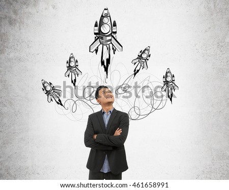 Asian businessman in suit is standing in front of concrete wall thinking about solution to difficult problem and smiling. Sketch of rockets on background. Concept of decision making.