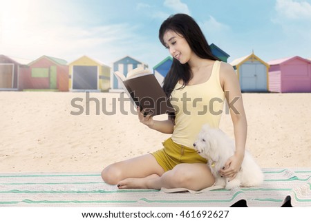 Portrait of a beautiful female model sitting on the beach while reading a book with her dog