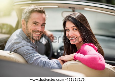 Rear view, looking at camera a cheerful couple in their convertible car, the woman is brunette and the man has grey hair. Shot with flare