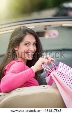Looking at camera, a cheerful brunette woman sitting in her convertible car and depositing shopping bags on the back seat. Shot with flare