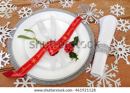 Christmas dinner decorative table setting with white porcelain plates, red ribbon, cutlery, linen napkin, holly, mistletoe and silver snowflake decorations over oak background.