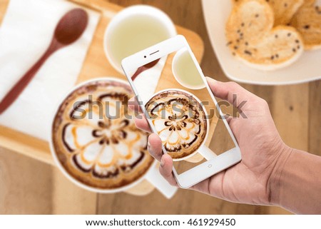 Men hand holding and using mobile (smart phone) over blurred A cup of coffee with flower latte art on top and bread heart and hot pandan juice, Concepttake a picture with smartphone.