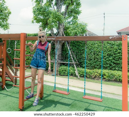 attractive young asian girl wear jean, sun glasses posting on modern kids playground  in green garden on summer day