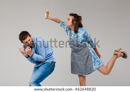 Young woman beating man with rolling pin over grey background.