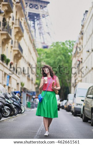 Cheerful young French woman walking with coffee to go and baguette (white bread) on a street of Paris, Eiffel tower in the background