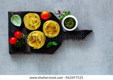 Traditional Italian cooking - pasta tagliatelle, pesto sauce, cherry tomatoes and basil on dark rustic wooden cutting board over light grey background, top view, copy space