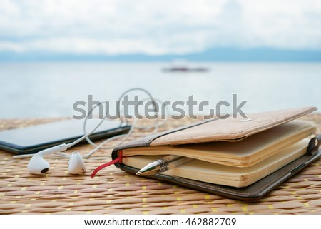 Closed notepad, pen and phone with headphones against tropical sea under cloudy sky