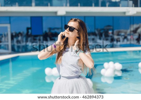 Young pretty girl with long hair and in black sunglasses is standing near pool. She wears gray T-shirt and skirt. She is happy speaking on phone.