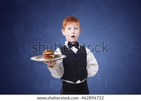 Little surprised waiter stands with tray serving hamburger. Redhead child boy in suit plays restaurant servant, gives burger at blue background