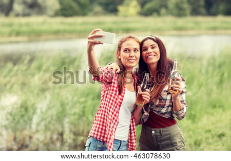 camping, travel, tourism, hike and people concept - happy young women with glass bottles drinking cider or beer and taking selfie by smartphone outdoors