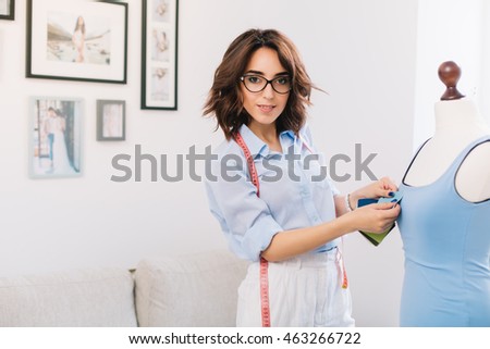 A brunette girl in a blue shirt is making  dress in the workshop studio. She has sewing stuff in hands. She is looking to the camera.