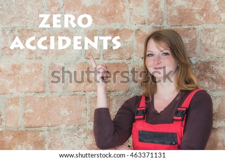 Young woman dressed in red overall is standing  in front of an old brick wall. Woman is looking at the camera and is pointing the finger at the sign Zero Accidents. 