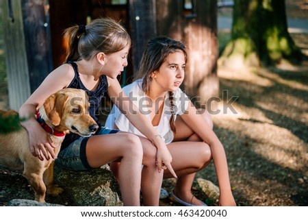 Two talking girls on a playground in summer. They have a small yellow dog. 