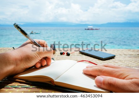 caucasian man is writing sime idea, message or letter in his notepad by pen while he sitting on the beach of tropical sea with boat