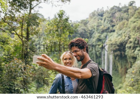 Happy young couple taking self portrait with their cell phone on hiking day. Couple taking picture in front of waterfall.