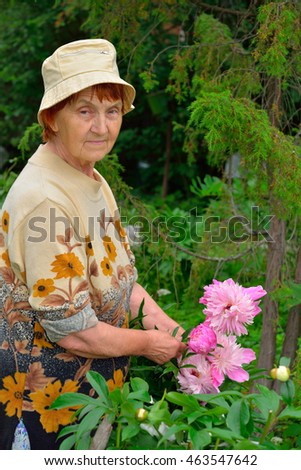 An elderly woman gardener standing and smiling next to pink peonies summer in Leningrad region