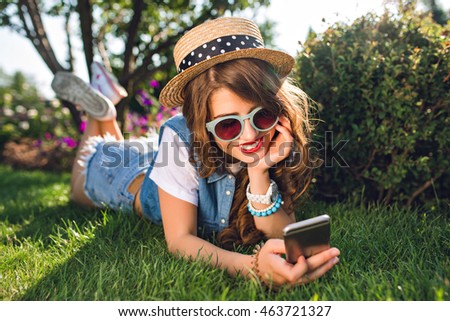 Portrait of cute  girl with long curly hair in hat lying on grass in summer park. She wears jeans jerkin,  shorts, sunglasses. She is typing on phone.