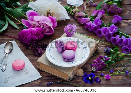Purple and pink French macaroon cookies on a white plate on a book and a variety of pink and purple flowers on a wooden background