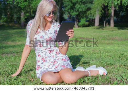 Girl sitting on the grass in the park among the trees and use the Internet on your tablet. Wireless internet connection. Social networking and chat for mobile communication.