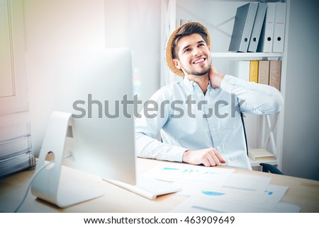 Happy relaxed young businessman in hat sitting and dreaming at workplace